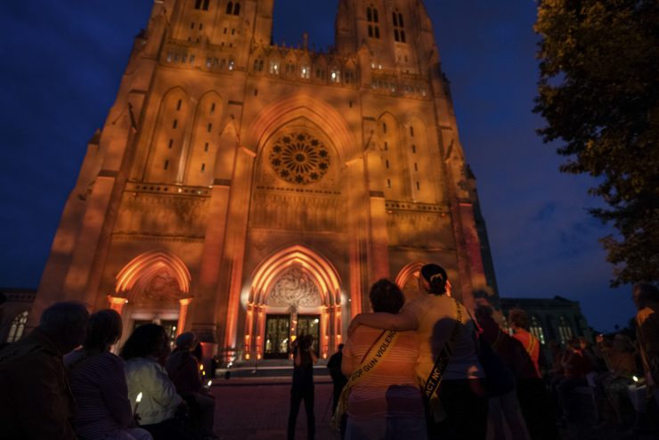 Anti-gun violence activists hold a vigil outside an orange-lit Cathedral at dark.