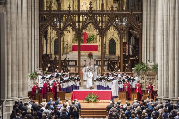 Acolytes, clergy, and choir members stand on a large platform in front of a congregation.